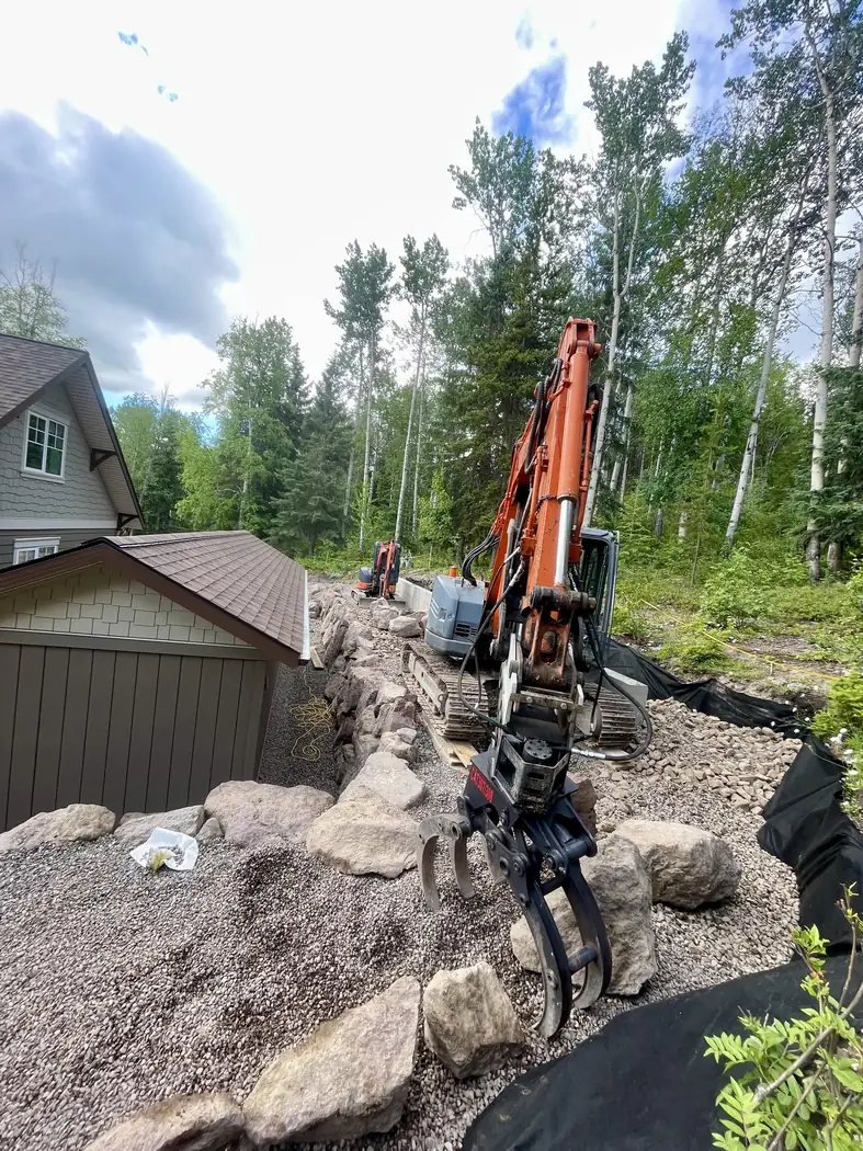 Excavator with a grapple setting boulders into a rock retaining wall beside a home